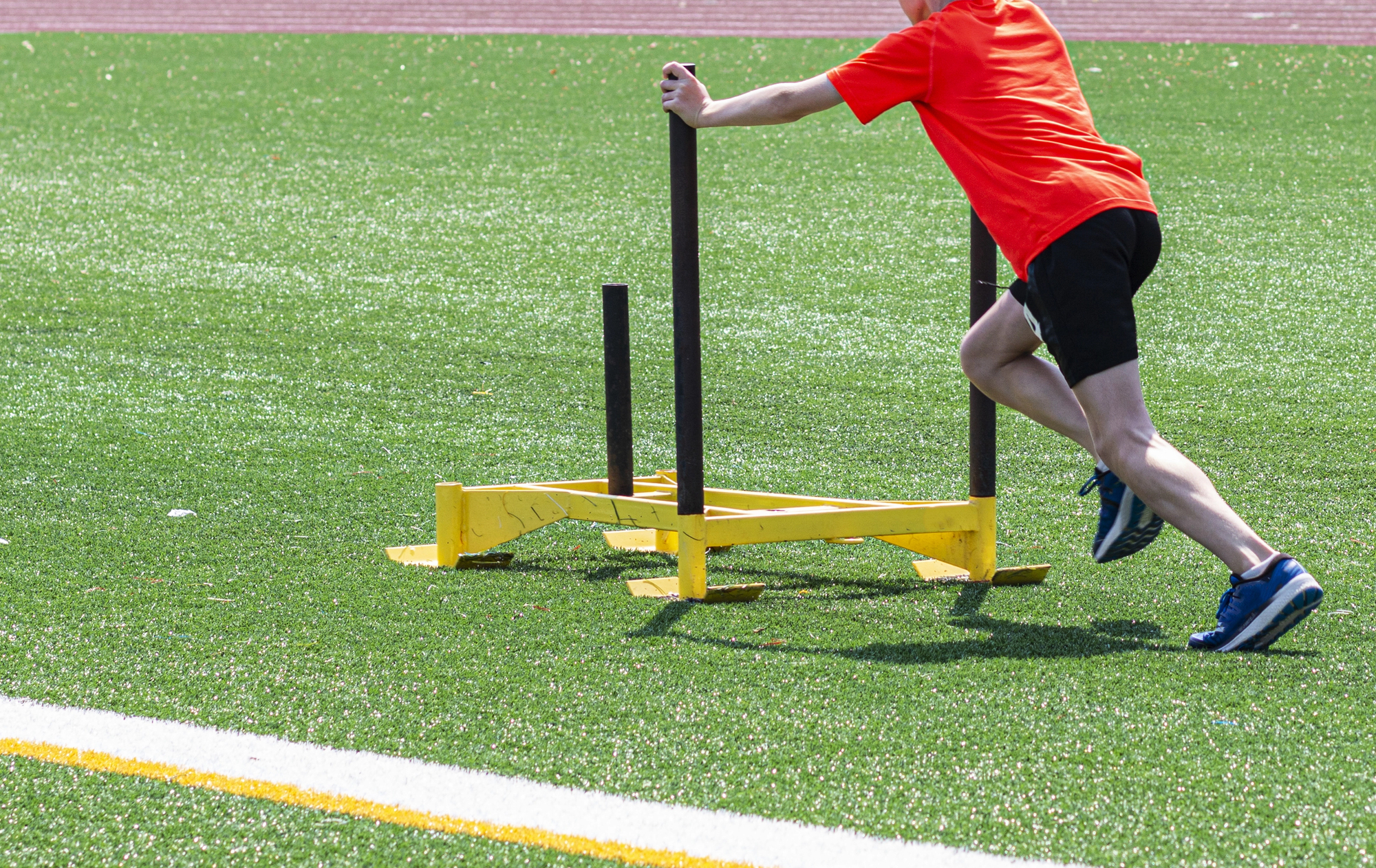 A young male runner is pushing a yellow sled on a green turf field at running camp. — Photo by WoodysPhotos